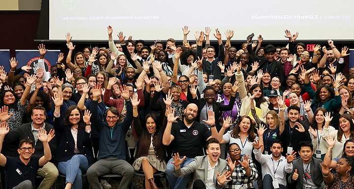 NSLS Summit attendees packed together in rows with their hands raised in the air.