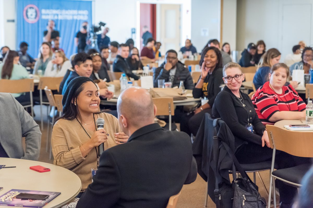 NSLS Summit attendees seated at round banquet tables in a session room. One attendee holds a microphone mid-sentence, facing a facilitator seated in the foreground, while people at nearby tables listen.